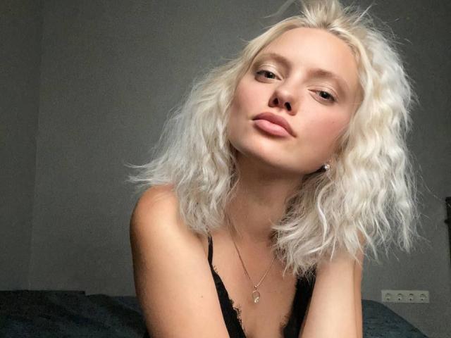 Close-up photo of CrystalHope, a blonde woman with messy hair, wearing a black tank top, leaning forward with a seductive gaze, light skin, slightly parted lips, visible necklace. Sexual tension.