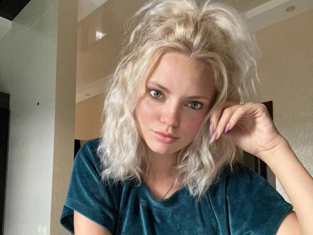 Close-up photo of CrystalHope, a blonde woman with messy hair, wearing a black t-shirt, blue eyes, and long purple nails, looking seductively at the camera with a slight smirk.