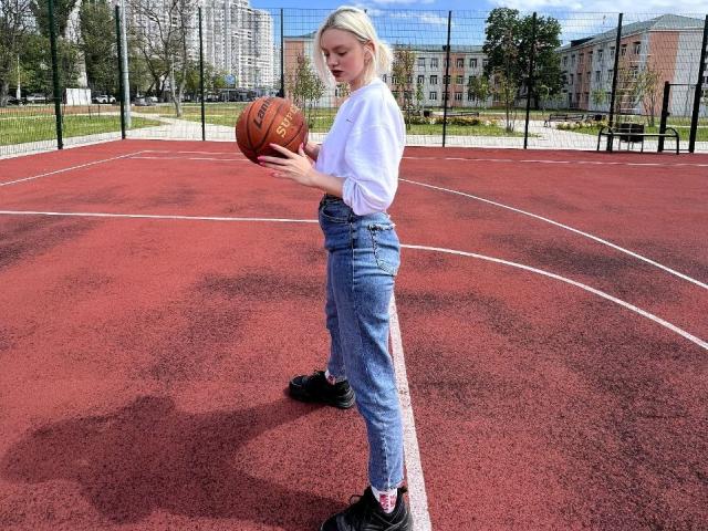 CrystalHope, eine blonde Frau, steht auf einem roten Basketballplatz und hält einen Basketball. Sie trägt ein weißes Hemd, blaue Jeans und schwarze Turnschuhe, im Hintergrund sind städtische Gebäude zu sehen. Verdammt heiß.