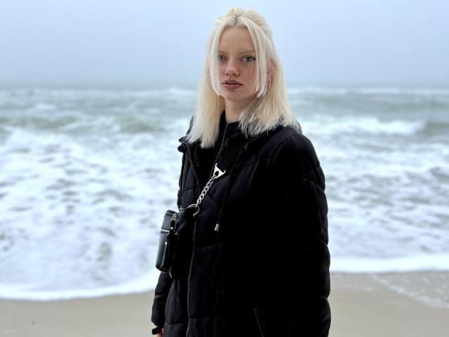 Foto van een blonde vrouw die op een strand staat, gekleed in een zwarte jas, met een camera om haar nek. Ze heeft een serieuze uitdrukking, met op de achtergrond golven van de oceaan. Het is verschrikkelijk koud, lijkt me..