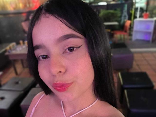 Primo piano di LissyCarter, una giovane donna con lunghi capelli neri, pelle chiara e rossetto rosso, che fa un leggero labbro. Indossa una canotta rosa a spalline sottili. Lo sfondo mostra mobili sfocati e luci al neon..