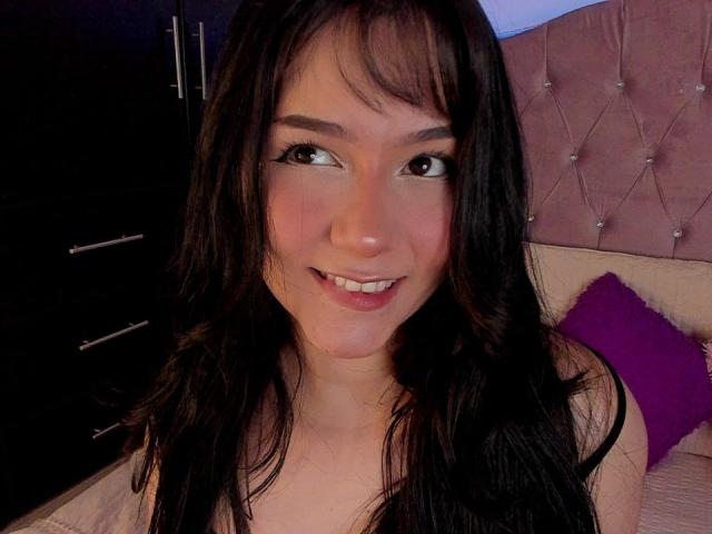 Nahaufnahme von DeisyWest mit langen, schwarzen Haaren, die in die Kamera lächelt. Sie hat dunkle Augen und helle Haut. Ein rosa, mit Fransen versehenes Kopfteil und ein violettes Kissen sind im Hintergrund zu sehen. Sexy, leicht verführerischer Look.