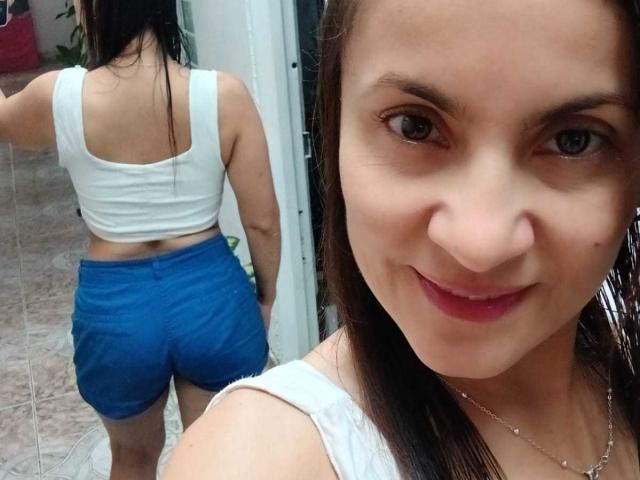 Close-up photo of SolaraDubois, brown-eyed woman with straight black hair, white tank top, and blue shorts, smiling at camera; background shows SolaraDubois from behind, same outfit, tiled floor.