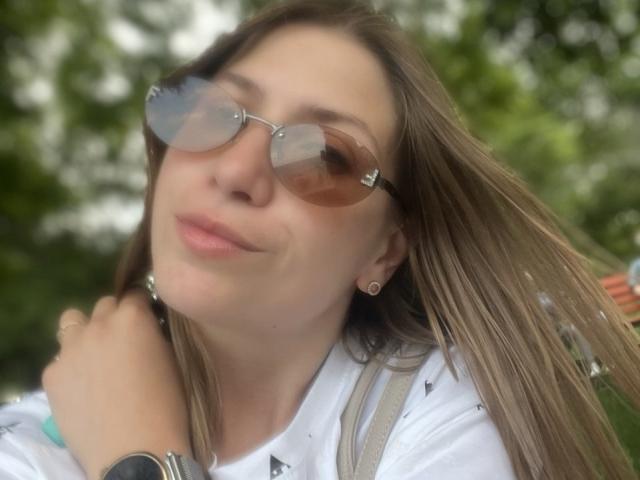 Close-up de LoriEla com cabelos castanhos longos, usando óculos de sol estilo aviador e uma camisa branca, com a mão no ombro, um leve sorriso. Fundo verde desfocado, casual, sexy, pose relaxada, ambiente externo..