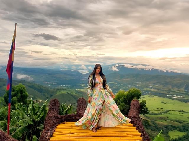 CatalinaRivero stands on a yellow wooden bridge, wearing a floral dress, with long black hair. Background: lush green hills, cloudy sky, colorful flag on the left. Fucking gorgeous view.