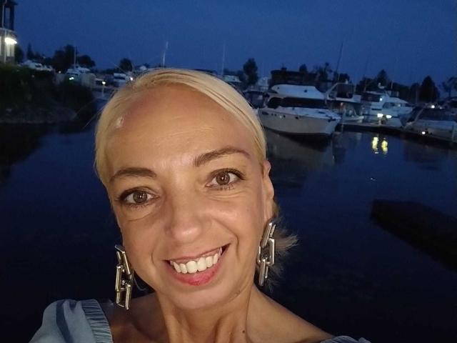 Fotografia em close-up de LunaBriella, uma mulher sorridente com cabelo loiro curto, usando brincos geométricos, em um fundo escuro. Fundo de marina azul ao entardecer. Barcos e janelas iluminadas ao fundo. Um sorriso incrivelmente atraente..
