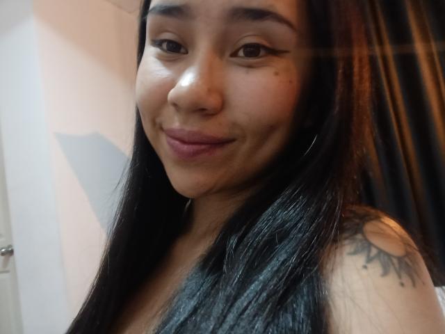 Primer plano de Karollteyes con cabello negro largo, ojos marrones y una sonrisa sutil. Ella tiene un tatuaje de un sol en el hombro derecho y lleva una camiseta oscura sin mangas. Muy atractivo/a..