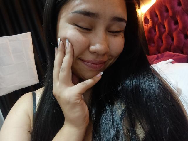 Close-up photo of Karollteyes, a woman with long black hair, eyes closed, smiling softly, touching her cheek with her right hand. Background includes red tufted headboard and white bed. Sexual tension.