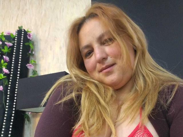 RouseWright heeft blond, lichtelijk rommelig haar, een lichte huid en een subtiele glimlach. Ze draagt een rode kanten bh en een donkere cardigan, met paarse bloemen op de onscherpe achtergrond. Heel aantrekkelijk..
