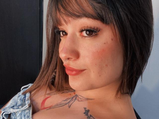 Close-up of AnikaFleur, a fair-skinned woman with brown hair and bangs, looking over her shoulder. She has a nose ring, tattoos on her neck and chest, wearing a denim shirt. Her lips are glossy.