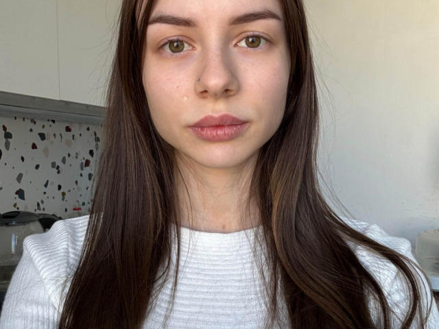 Gros plan de Lavenia Wimes, une jeune femme aux longs cheveux bruns lisses, portant un t-shirt blanc côtelé. Son expression est neutre, avec des lèvres pulpeuses et une peau légèrement rosée. Extrêmement sexy..