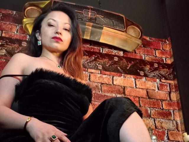 Photographie d'Angelina Blanche avec des cheveux foncés, du rouge à lèvres rouge et une robe en fourrure noire sans bretelles, assise devant un mur de briques rouges. mur avec un emblème de voiture jaune et noir au-dessus. Pose incroyablement séduisante..