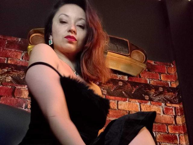 Fotografie von Angelina Blanche mit rotem Lippenstift, einem schwarzen, trägerlosen Kleid und dunkelbraunen Haaren, die nach unten in die Kamera blickt. Ziegelwand im Hintergrund, gelüfteter Lüftungsgitter über ihrem Kopf, verführerische Pose..