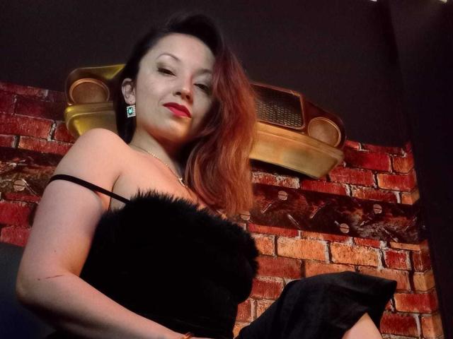 Fotografía de Angelina Blanche con cabello rojo, vestida con un vestido negro sin tirantes con detalles de piel, sentada contra un muro de ladrillos. pared. Ella tiene labial rojo, aretes oscuros y una sonrisa seductora..