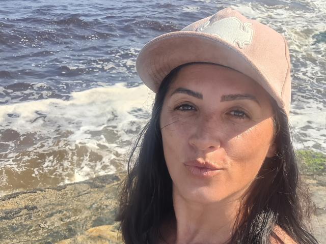 Photograph of RenataAlves, a woman with long black hair, wearing a beige hat, standing by a sunlit ocean. Her expression is neutral, sunlight highlights her face, water splashes behind her.