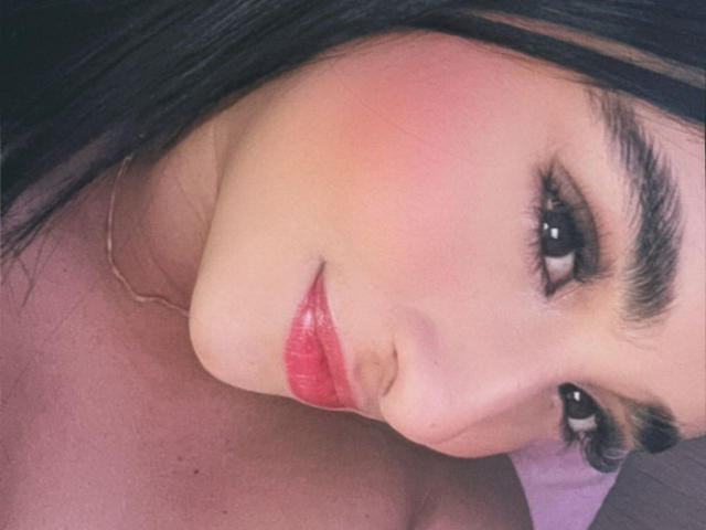 Close-up foto van EmmaWaltter, met de focus op haar gezicht en bovenborst. Ze heeft donker haar, zwarte wenkbrauwen en felrode lippenstift. Haar huid is bleek met een lichte bloos. Ze kijkt verleidelijk in de camera met een subtiele glimlach..