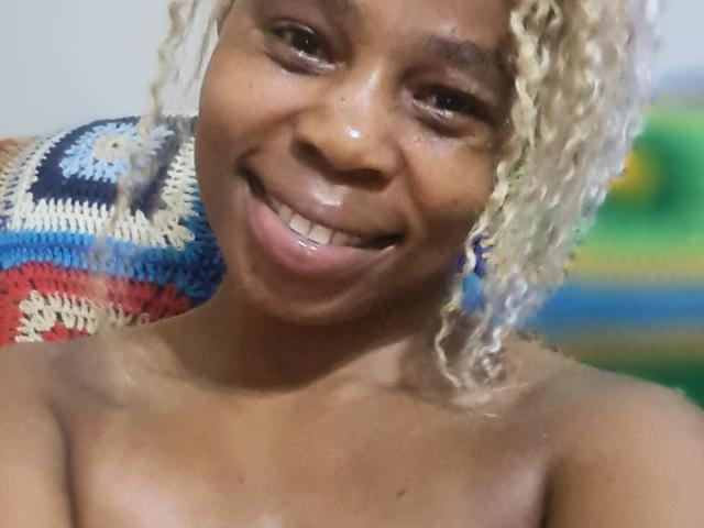 Close-up de um traseiro atraente, sorrindo com lábios brilhantes, pele marrom escura e cabelo loiro cacheado. O fundo apresenta um cobertor de tricô colorido e tons verdes e azuis desfocados. Focado na expressão facial.