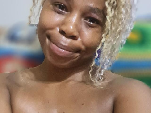 Close-up of SexyHotButt, smiling with glossy skin, light curly blonde hair, dark brown eyes, topless, blurred colorful background, visible droplets on face, slight tilt, casual, intimate, suggestive.