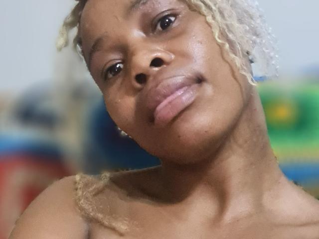 Close-up photo of SexyHotButt, dark-skinned woman with light blonde braids, glossy lips, and a subtle smile. Blurry background with green and blue hues. Focus on face, slightly tilted head.