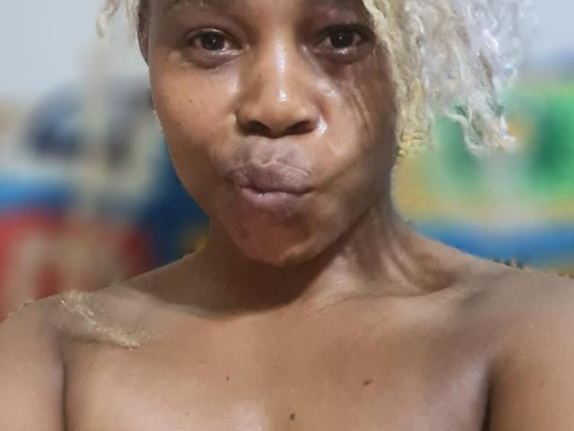 Close-up photo of SexyHotButt, dark-skinned with shiny, sweaty face and light blonde curls, pouting lips, bare shoulders, blurred colorful background. Foreground focus on glistening skin and intense gaze.