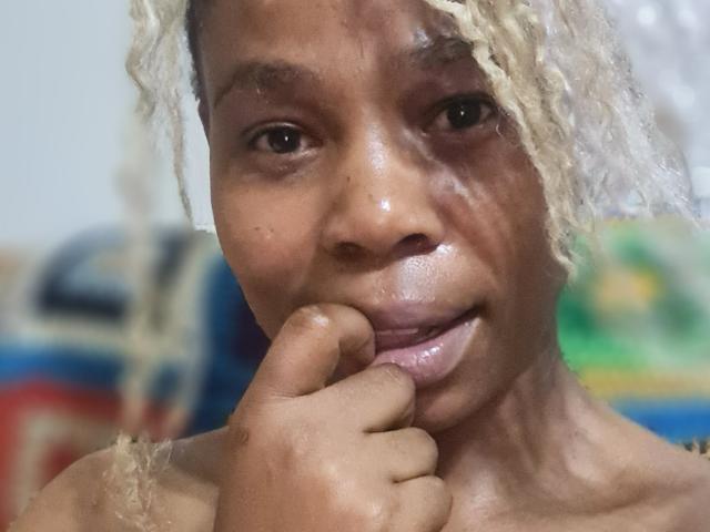 Close-up of SexyHotButt with dark brown skin, wet and shiny face, biting finger, blonde curly hair, intense gaze. Background blurry with colorful shapes. Slight sexual tension from provocative pose.