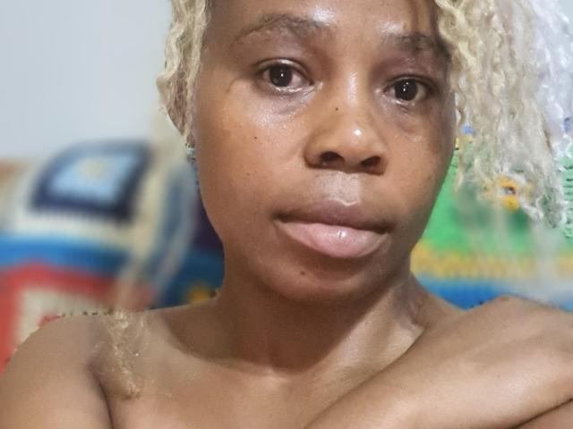 Close-up of SexyHotButt, a dark-skinned woman with curly blonde hair, glistening skin, and a neutral expression. Background is blurred with colorful shapes. Focused on face, shoulders, and intense gaze.