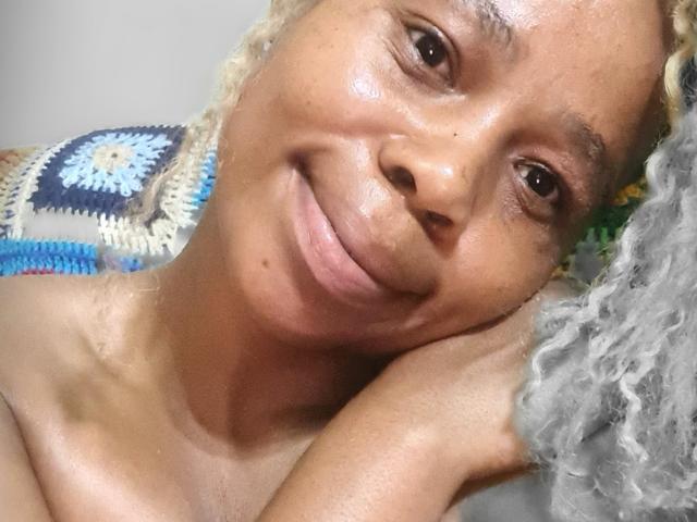 Close-up photo of SexyHotButt, a smiling black woman with curly blonde hair, resting her head on her hand. She has a soft, brown complexion, and wears a blue and white crocheted top.