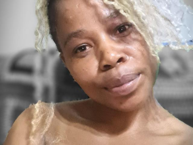 Close-up photograph of SexyHotButt with light brown skin, short curly blonde hair, and a slight smile. Naked shoulders, slight sheen on face, blurry background. Sexual tension, casual pose.