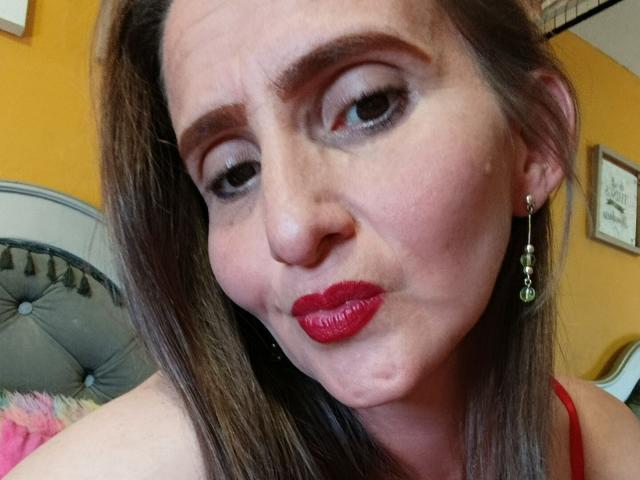 Close-up of RubiAnderson, a woman with pale skin, red lipstick, and dark brown hair, wearing red earrings and a red top. She has a slightly cocked head and a playful expression.