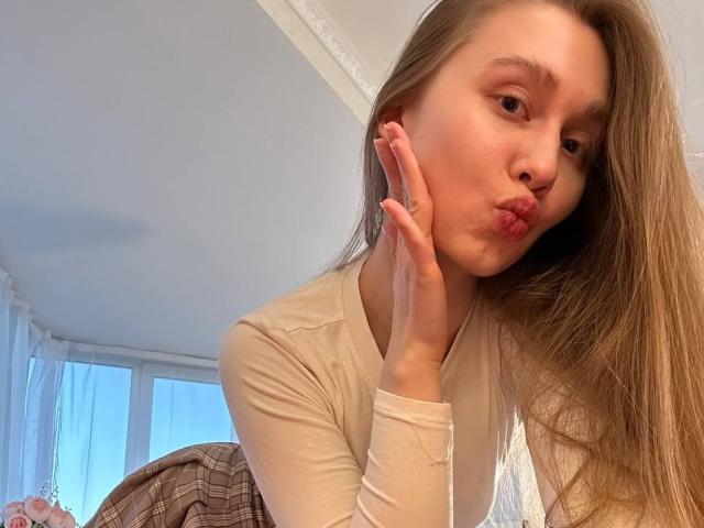 Close-up of KelseyCaswell with long brown hair, pouting lips, and hand on cheek, wearing a white long-sleeve top, leaning forward, bright sunlight from window, plaid blanket in background.