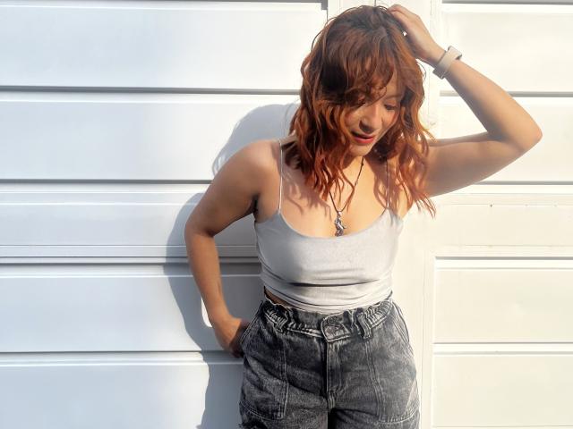 Photograph of EmilyKlein with wet, shoulder-length brown hair, leaning against a white wall. She wears a white tank top, high-waisted black jeans, and a necklace. Hand in pocket, head tilted, shadowed face.