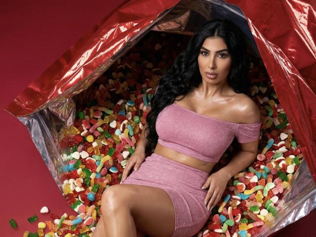 BrunetteBabe69, com cabelos longos e pretos, vestindo uma blusa cropped rosa com glitter e um mini saia combinando, sentada em um bolsa vermelha cheia de doces coloridos, olhando sedutoramente para a câmera.