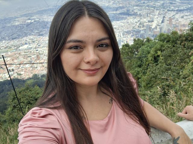 Fotografía de una mujer de origen latino con cabello largo y oscuro, vestida con una camisa rosa, sonriendo y de pie en un mirador de montaña. Paisaje urbano en primer plano, vegetación densa en el fondo. Expresión enfocada, ligeramente seductora..