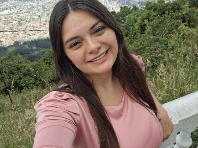 Photograph of a young woman with long dark hair, wearing a pink shirt, smiling at the camera. She has a visible nose piercing and is leaning on a white railing. Background includes green trees and a distant cityscape. Her breasts are prominent. Slight sexual tension from her confident expression.