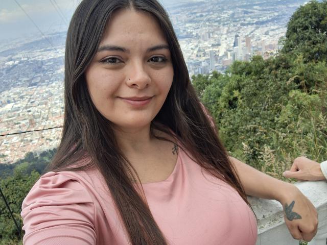 KatherinLatin, com longos cabelos pretos e uma blusa rosa, está apoiada em um corrimão, olhando para a câmera. Ela tem uma tatuagem de borboleta no pulso direito. Uma paisagem urbana incrivelmente sexy ao fundo dela..