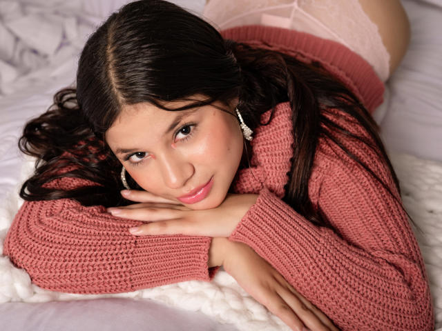 MilleyAngel est allongée sur un lit, la tête reposant sur ses mains, vêtue d'un pull rose en tricot et d'une culotte en dentelle blanche. Ses cheveux noirs encadrent son visage, et elle regarde la caméra de manière séduisante..