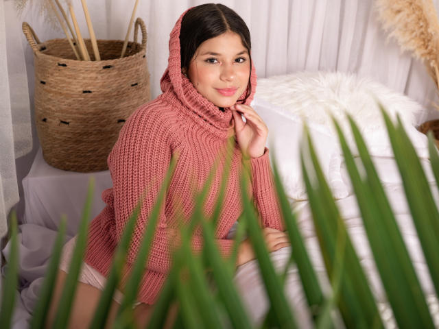 MilleyAngel with dark hair, wearing a red knitted hoodie, sitting on a bed with white sheets. Green leaves in the foreground, basket with reeds in the background. Fucking sexy.