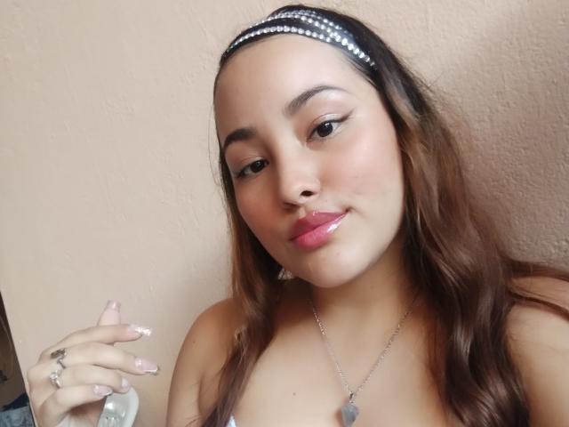 Close-up de IvannaVegaX, uma jovem com cabelos castanhos longos, usando uma faixa de cabeça preta e um colar de prata com um coração. pingente. Ela está segurando um preservativo, olhando diretamente para a câmera com um leve sorriso..