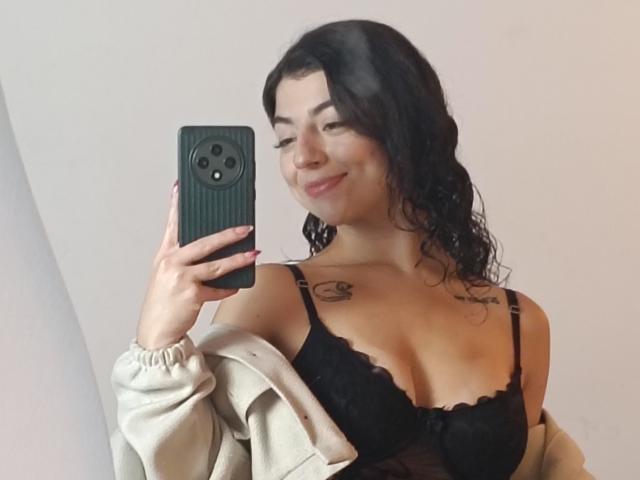 Anntonella Watson, met nat zwart haar, lachend, met een zwarte telefoon in haar hand, draagt een zwarte kanten BH en een beige jas die ze heeft uitgetrokken. schouders, tatoeages op de borst, zichtbare decolleté, selfie, casual, sexy.