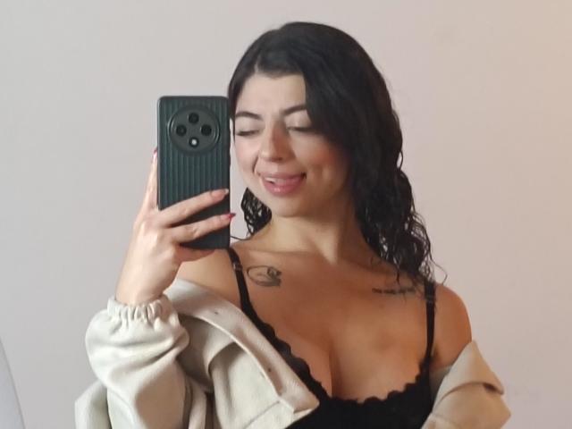 Anntonella Watson, con el cabello negro y mojado, sonriendo, sosteniendo un teléfono negro, vistiendo un sujetador de encaje negro y una chaqueta beige, con un tatuaje. en la clavícula derecha, busto medio, selfie casual, ligera tensión sexual..