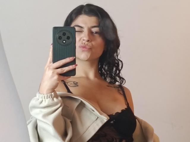 Selfie von Anntonella Watson mit nassen, welligen schwarzen Haaren, geschürzten Lippen, die ein schwarzes Telefon hält und eine beige Jacke trägt. Ein Schulterteil, schwarzer Spitzen-BH, kleines Tattoo an der rechten Schulter..