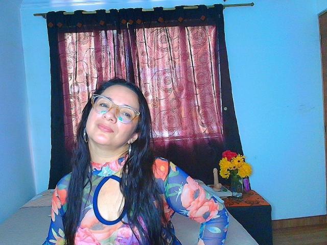 VictoriaRousse, long dark hair, wearing glasses and a floral top with a large circular cutout, leaning back with a suggestive smile. Background: blue walls, dark red curtains, flower vase. Bright lighting.