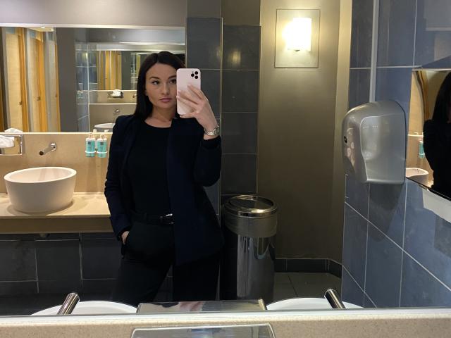 Dionisia, avec ses cheveux noirs et son rouge à lèvres rouge, portant une tenue noire, prend un selfie devant un miroir dans une salle de bain moderne. Elle tient un téléphone blanc et se tient près d'un évier blanc. L'éclairage est très lumineux..