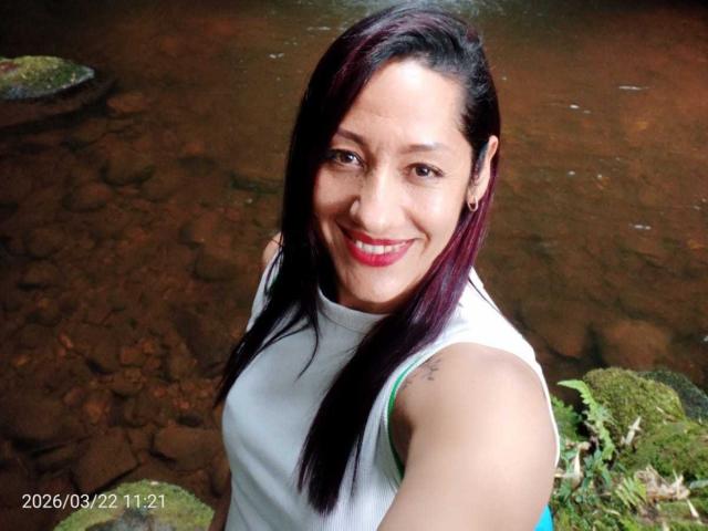 Fotografie von SamanthaCorine mit langen, schwarzen Haaren, die lächelt und roten Lippenstift trägt, und die ein weißes Top ohne Ärmel trägt. Im Hintergrund sind felsiger Boden und Grünflächen zu sehen. Zeitstempel: 22.03.2020 11:21. Sexuelle Anspannung angedeutet..