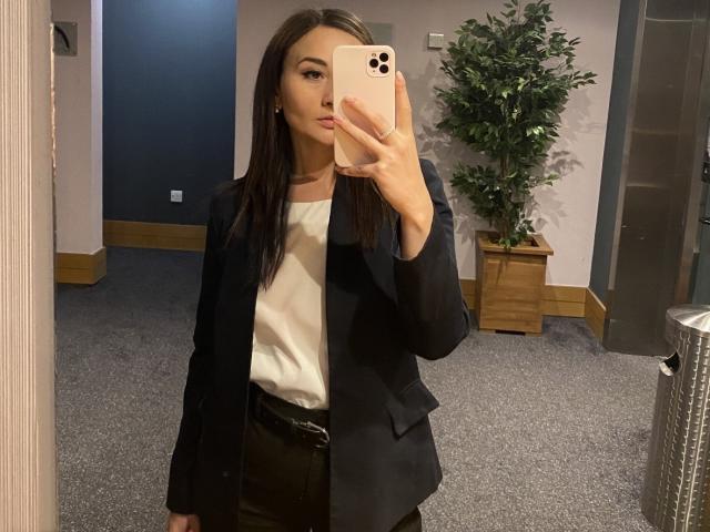 Dionisia está frente a un espejo, sosteniendo un iPhone dorado, y se está tomando una selfie. Ella lleva un blazer negro sobre una camisa blanca, con pantalones oscuros. El fondo incluye una planta en una maceta. Su expresión es ligeramente afligida..