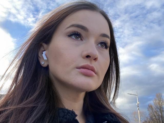 Close-up van Dionisia, een jonge vrouw met lang bruin haar, die een zwarte, pluizige jas draagt, met een blauwe lucht met wolken op de achtergrond. Achtergrond. Haar uitdrukking is serieus, met licht openstaande lippen. Helder daglicht.