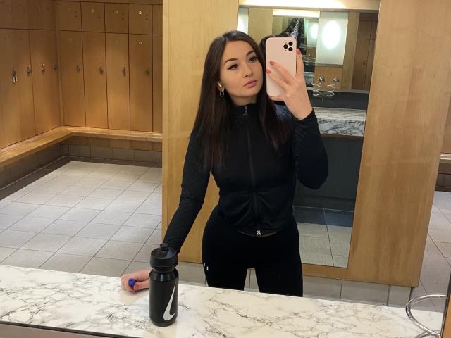 Dionisia è davanti a uno specchio e sta scattando un selfie con il suo iPhone. Indossa un abito nero aderente, e la sua mano è appoggiata su un bancone di marmo, mentre tiene una bottiglia nera. Specchio riflettente, armadietti in legno sullo sfondo.