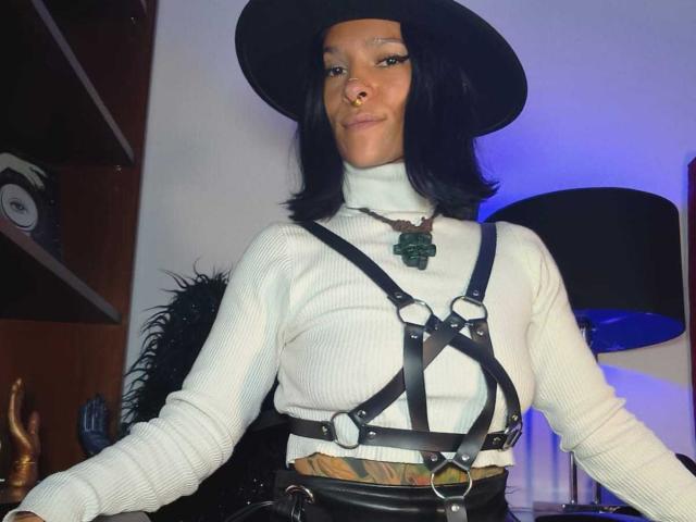 AmelieBlake wears a black hat and white ribbed turtleneck with black leather harness straps. She's smiling confidently, hands on table, with blue lighting in the background. Sexy, dominant vibe.