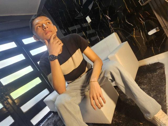 SamuJasper, sentada en un sofá blanco, vestida con un top corto negro y jeans azul claro, con la mano en la barbilla, mirando hacia arriba con una una expresión ligeramente arrogante. Pared de mármol negro con vetas en el fondo..