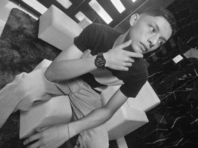 Fotografia in bianco e nero di SamuJasper, sdraiato su un divano, che indossa una maglietta nera e pantaloni beige, con un orologio al polso. dito sul mento, sguardo rivolto altrove, sensualità, stile moderno.